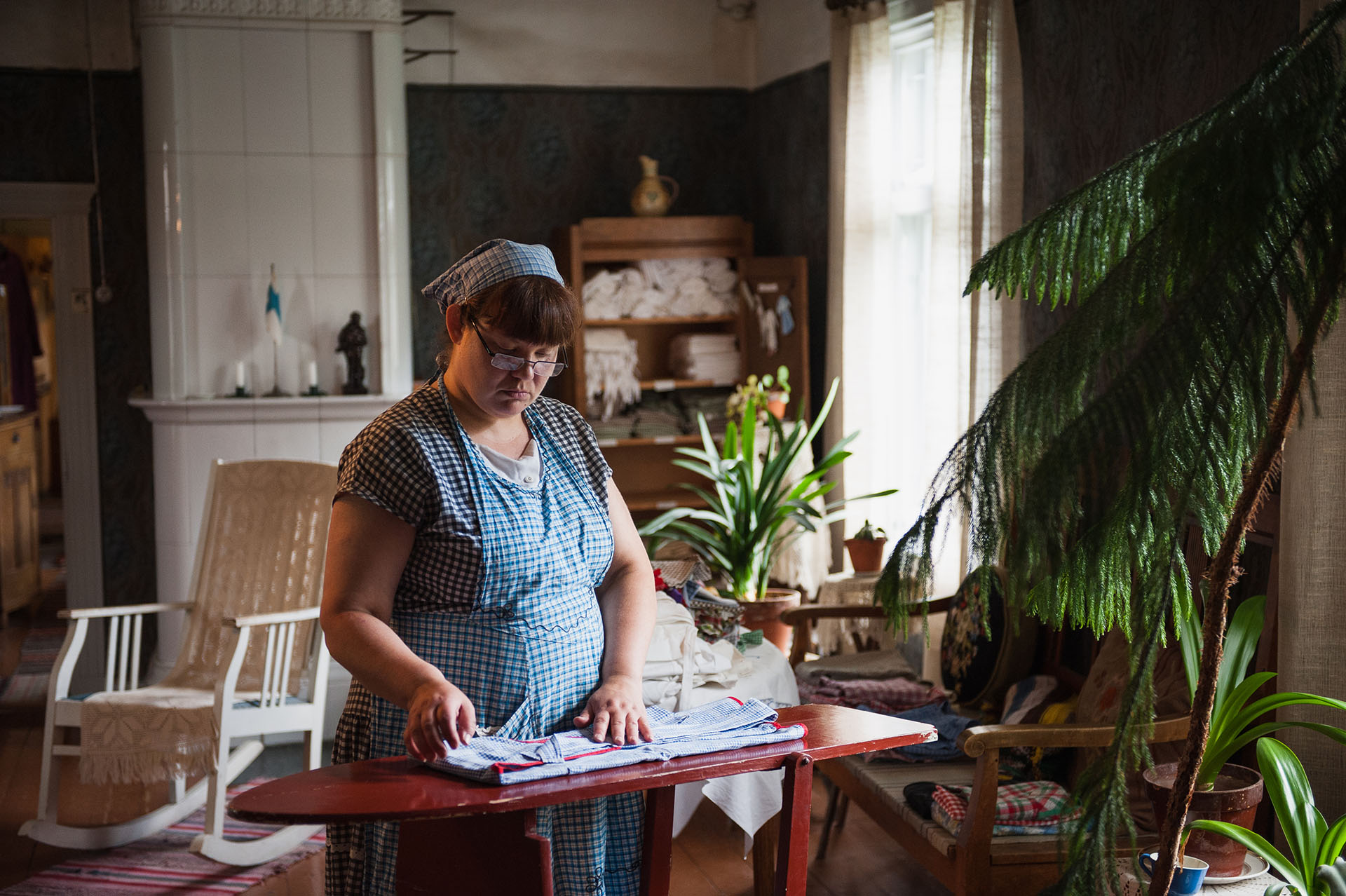 Ruudulliseen mekkoon, esiliinaan ja huiviin pukeutunut henkilö taittaa pyykkiä silityslaudalla. Taustalla salin kaakeliuuni, keinutuoli, avattu liinavaatekaappi ja huonekasveja.