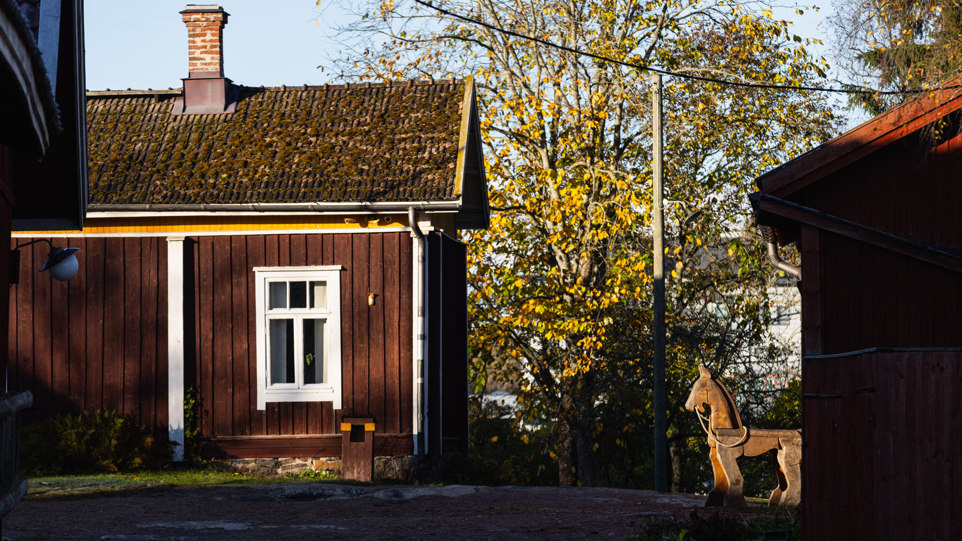 Punamullattu vanha rakennus, pihapiirissä iso puuhevonen, taustalla syyslehdissä oleva puu.